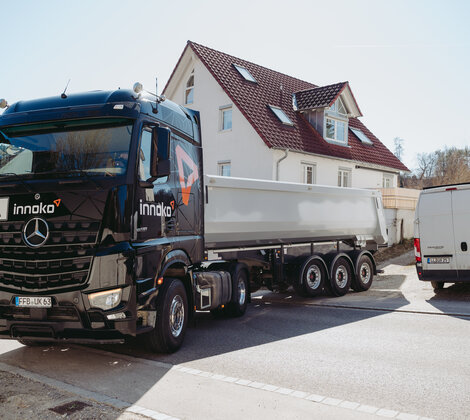 Schwarzer Mercedes-Benz-Lkw mit Auflieger der Firma Innoko parkt vor einem Wohnhaus mit rotem Dach; daneben steht ein weißer Transporte